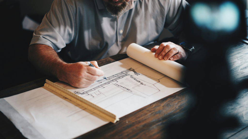 Un architecte dessine des plans de maison sur une feuille, avec une règle et un rouleau de papier, illustrant un projet immobilier.