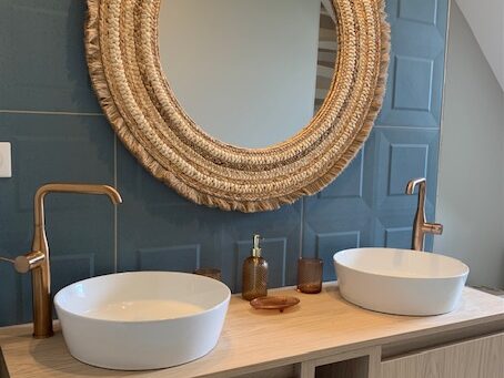 Salle de bains élégante avec deux vasques sur un meuble en bois, miroir tressé et robinetterie dorée.