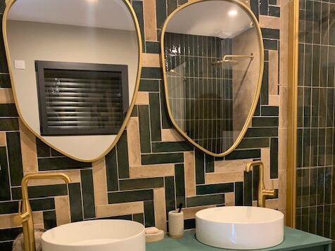 Salle de bains moderne avec deux lavabos ronds, miroirs ovales dorés et carrelage vert en motif chevron.